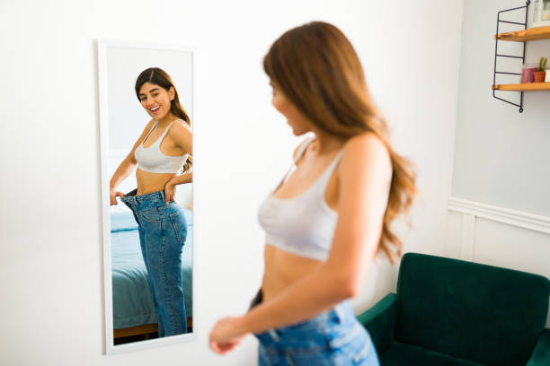 A woman admires herself in the mirror as she pulls the waistband of an oversized pair of jeans she's wearing away from her body